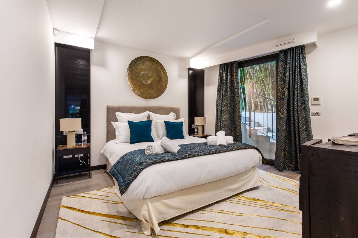 A modern hotel room with a large bed, a round wall decoration, a nightstand with a lamp, and a view of the outdoors through a window with curtains.