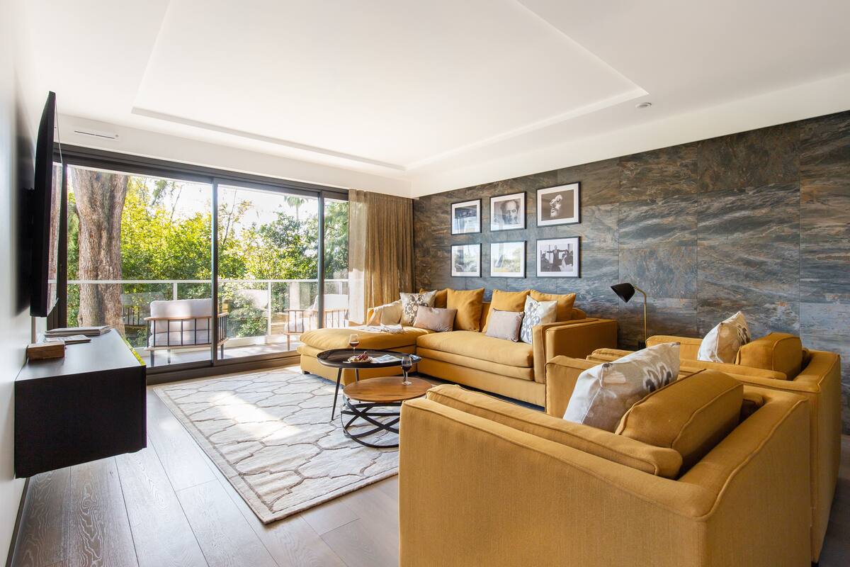 A modern living room with a large sectional sofa, a coffee table, and a flat-screen TV mounted on the wall. There is a large window providing a view of the outdoors.