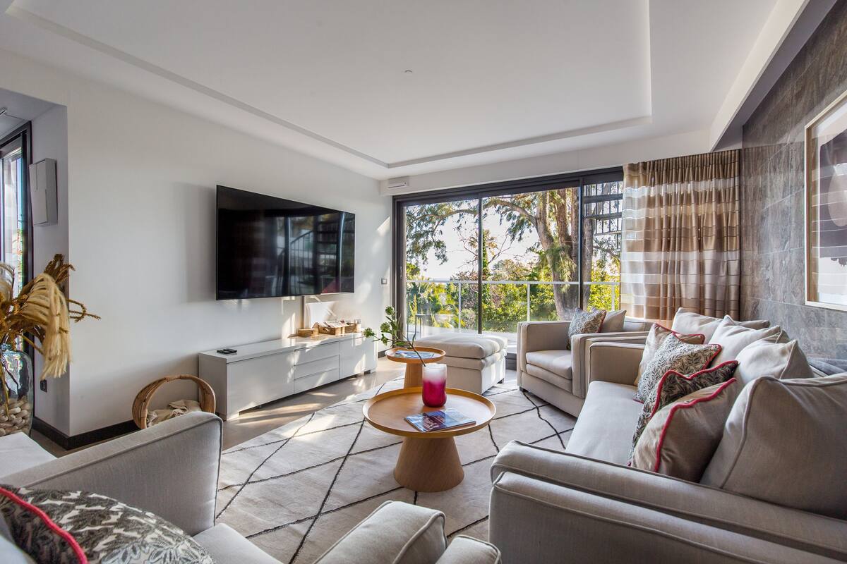 A modern living room with a flat-screen TV, comfortable seating, a coffee table, and a large window with curtains.