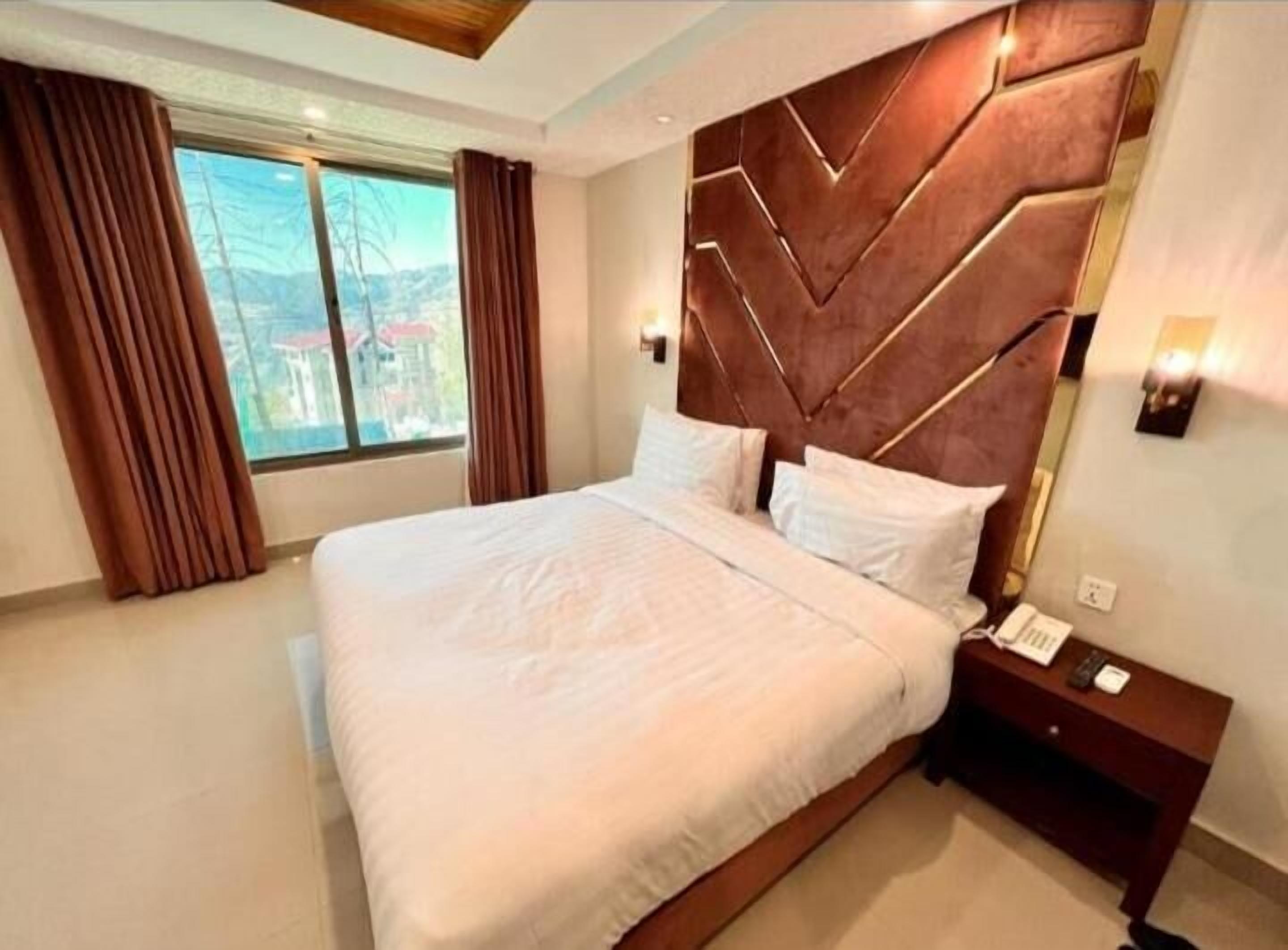 A modern hotel room with a large bed, a wooden headboard, a bedside table with a phone, and a view of the outdoors through the window.