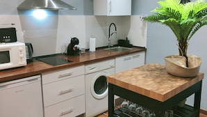 Cozinha compacta com micro-ondas, cafeteira, pia e máquina de lavar roupa. Há uma planta em vaso na bancada e uma mesa de madeira com uma cadeira.
