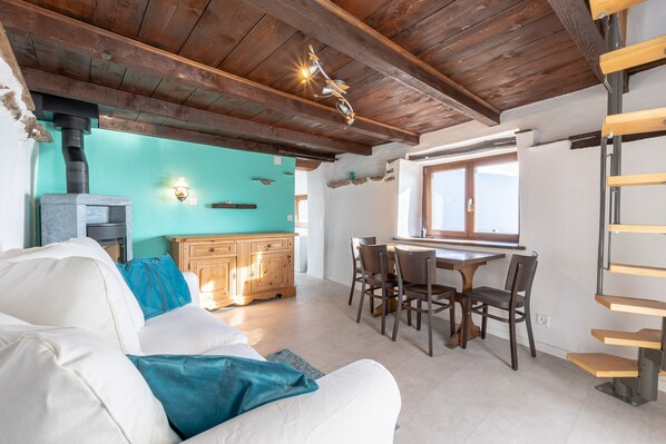 Un salon chaleureux avec un canapé blanc, des coussins turquoise, une table à manger en bois avec des chaises, et un coin cuisine équipé d’une cuisinière.