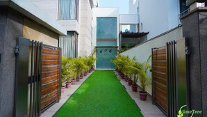 A narrow pathway with artificial green turf, flanked by potted plants and modern buildings with metal gates. - Lime Tree Hotel Sushant Lok Gurgaon  (Gurugram)