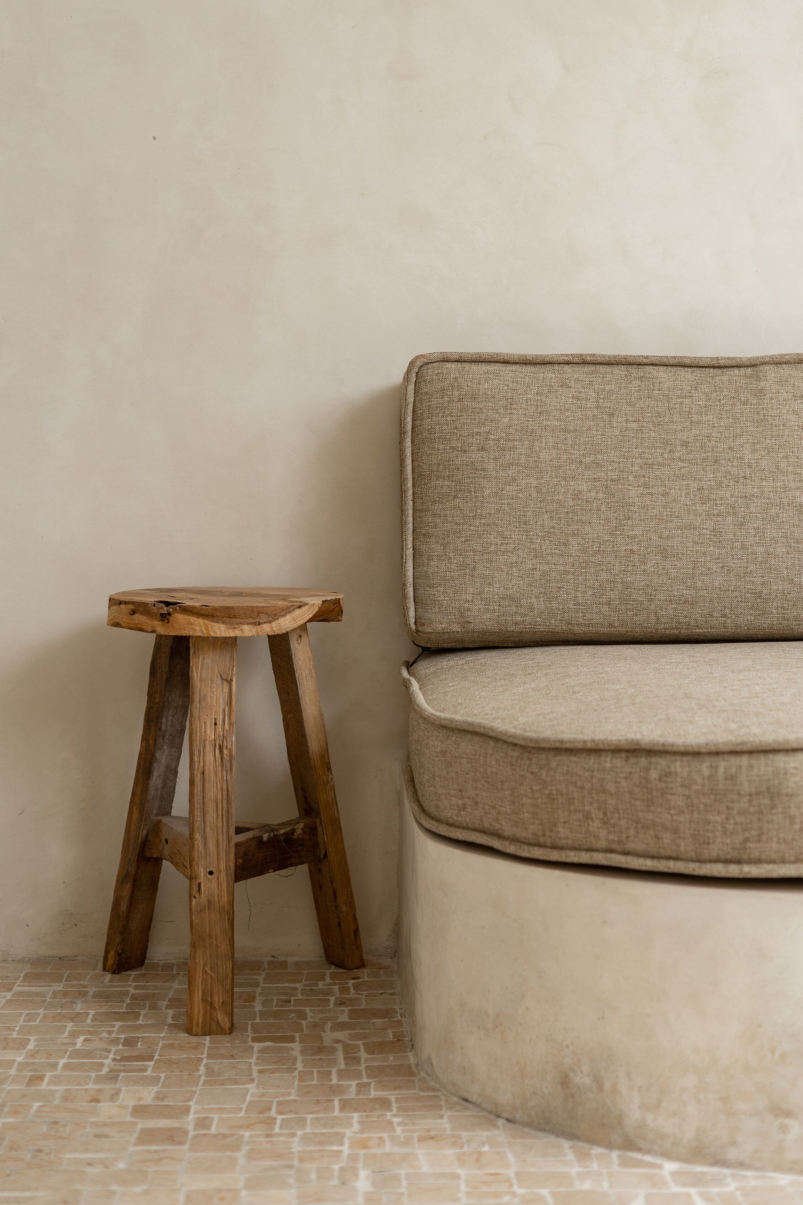 A corner of a room with a wooden stool and a beige sofa against a plain wall.