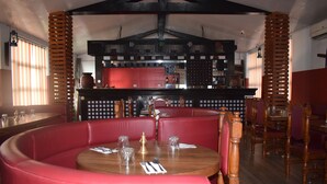 Un restaurante con zona de bar, asientos tapizados en rojo, mesas de madera y pared de ladrillo.