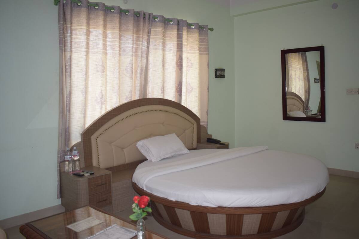 A bedroom with a large bed, a bedside table, a nightstand, a mirror, and a window with curtains.