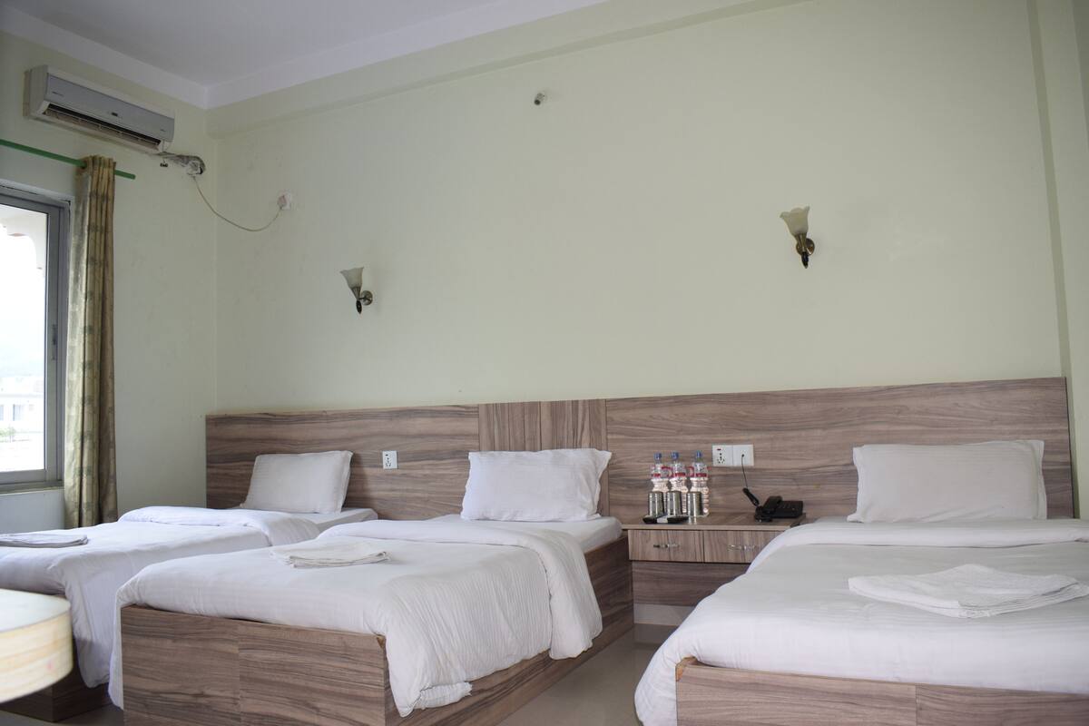 A hotel room with three beds, a window with curtains, an air conditioning unit, and a bedside table with a phone and bottled water.