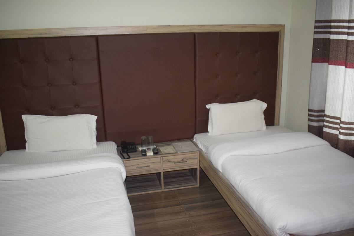 A hotel room with two single beds, a wooden nightstand with a phone and remote controls, and a brown headboard.