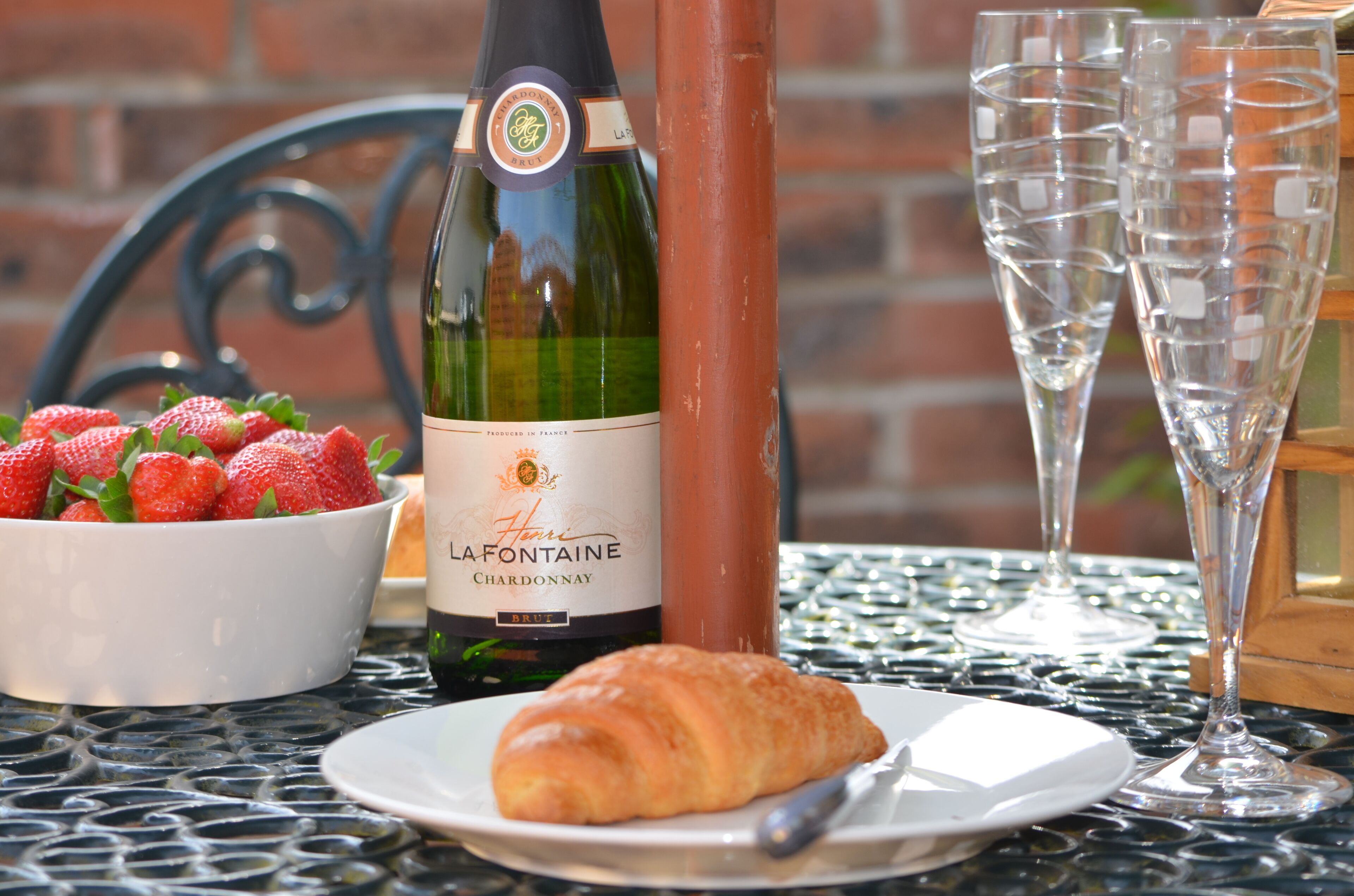 Eine Flasche La-Fontaine Chardonnay, eine Schale Erdbeeren, ein Croissant und zwei Sektgläser auf einer Metalltisch.