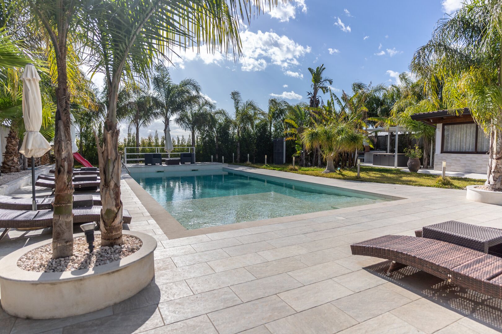 A swimming pool with lounge chairs and umbrellas, surrounded by palm trees and a modern building.