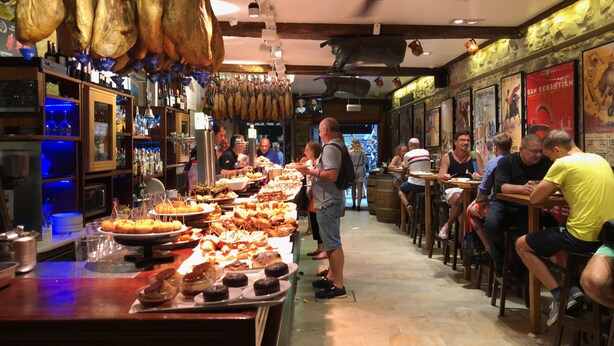A restaurant with a long counter filled with various dishes, a bar area with bottles, and hanging meats.