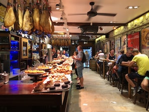 A restaurant with a long counter filled with various dishes, a bar area with bottles, and hanging meats.