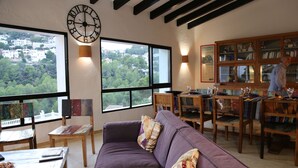 A living room with a purple sofa, wooden chairs, a dining table, and a large window with a view of a town and trees.