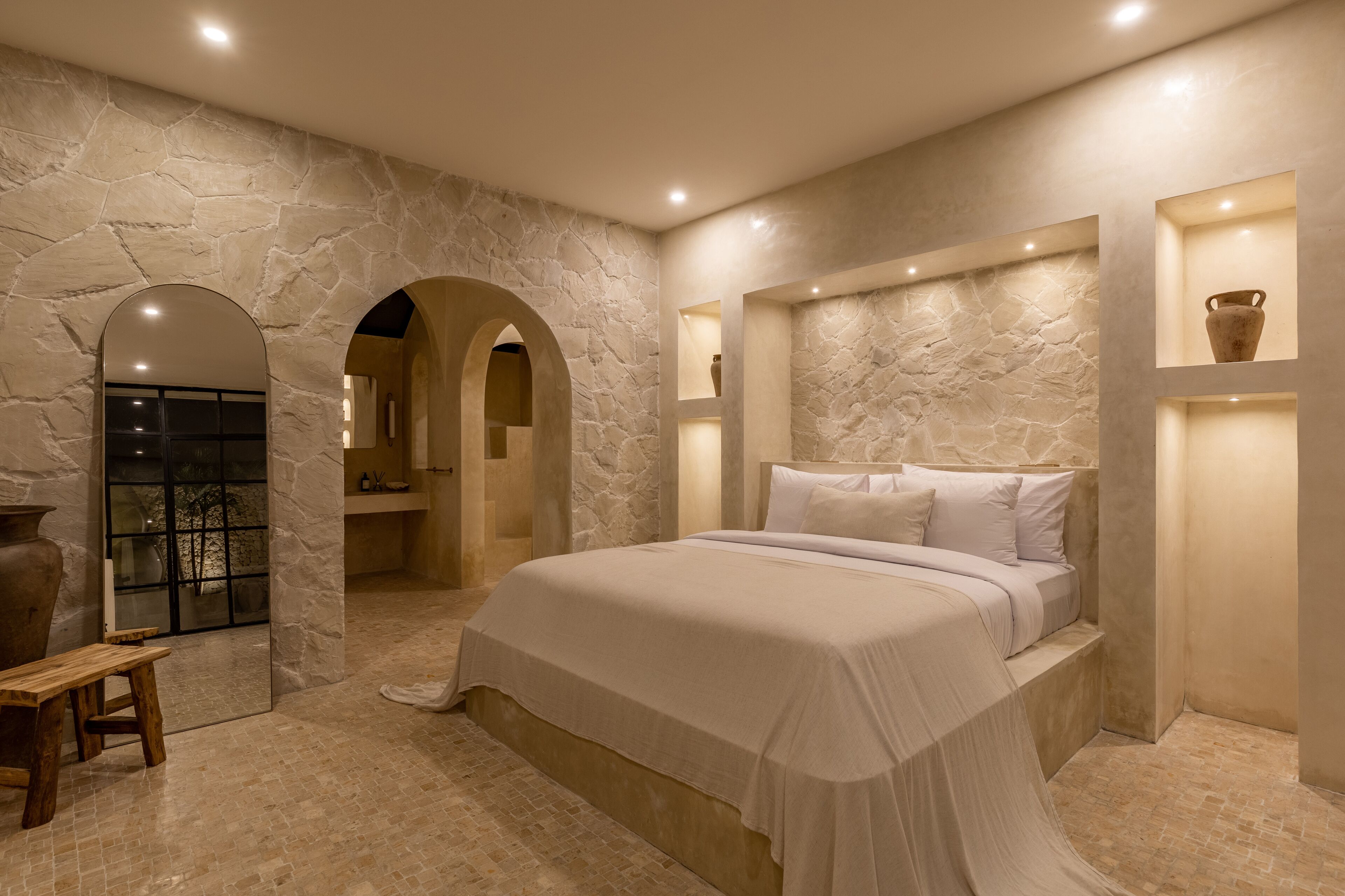 A bedroom with a stone wall, a bed, a bench, and a shelf with a vase.