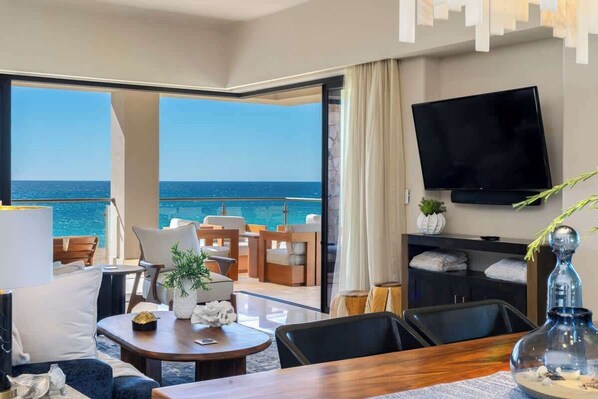 A modern hotel room with a balcony, a flat-screen TV, and a dining area with a view of the sea.
