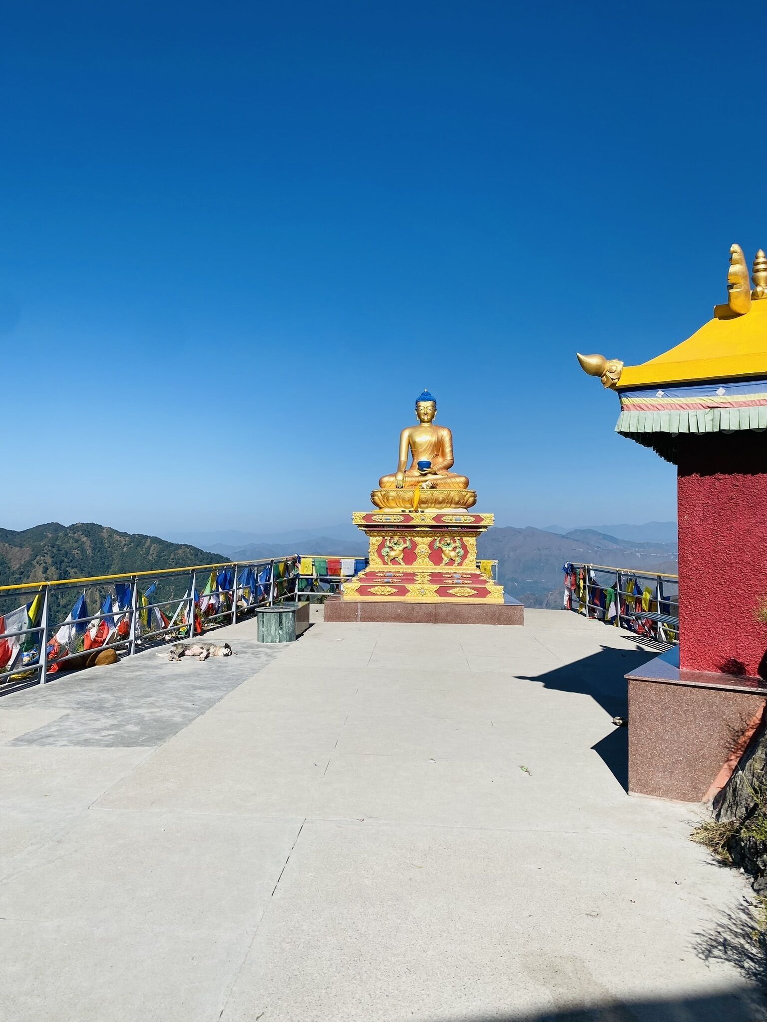 A golden Buddha statue on a pedestal with a red and yellow roof, surrounded by colorful prayer flags.