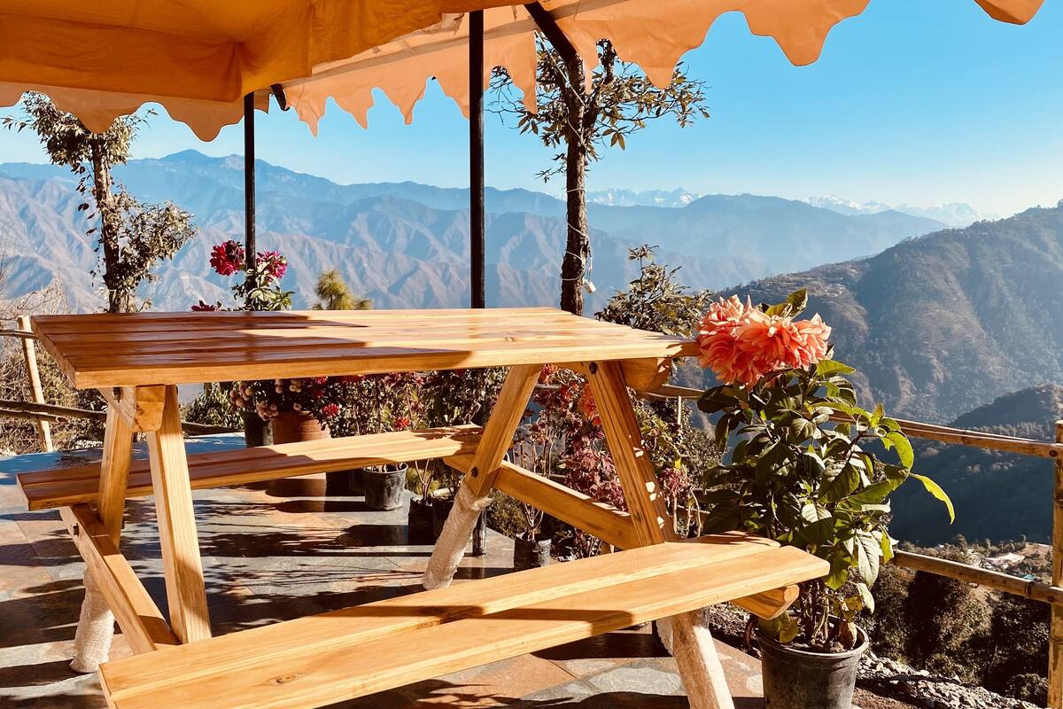 Une table et un banc en bois, placés sous une tonnelle, agrémentés de plantes en pot et offrant une vue sur les montagnes.