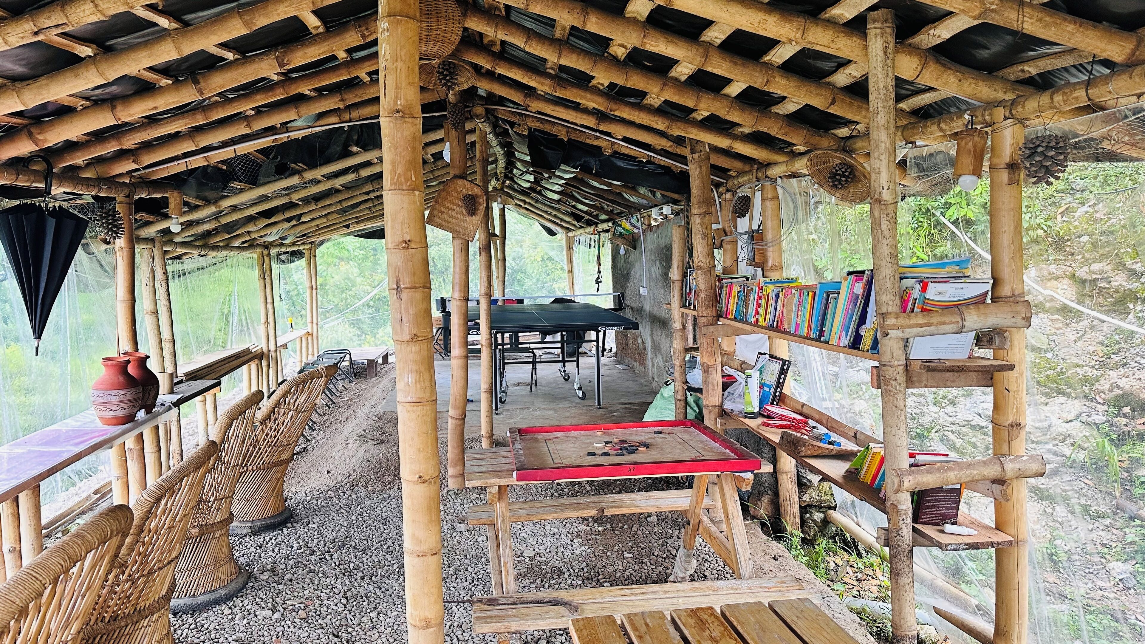 A bamboo structure with a table and chairs, a bookshelf with books, and a view of the surrounding greenery.