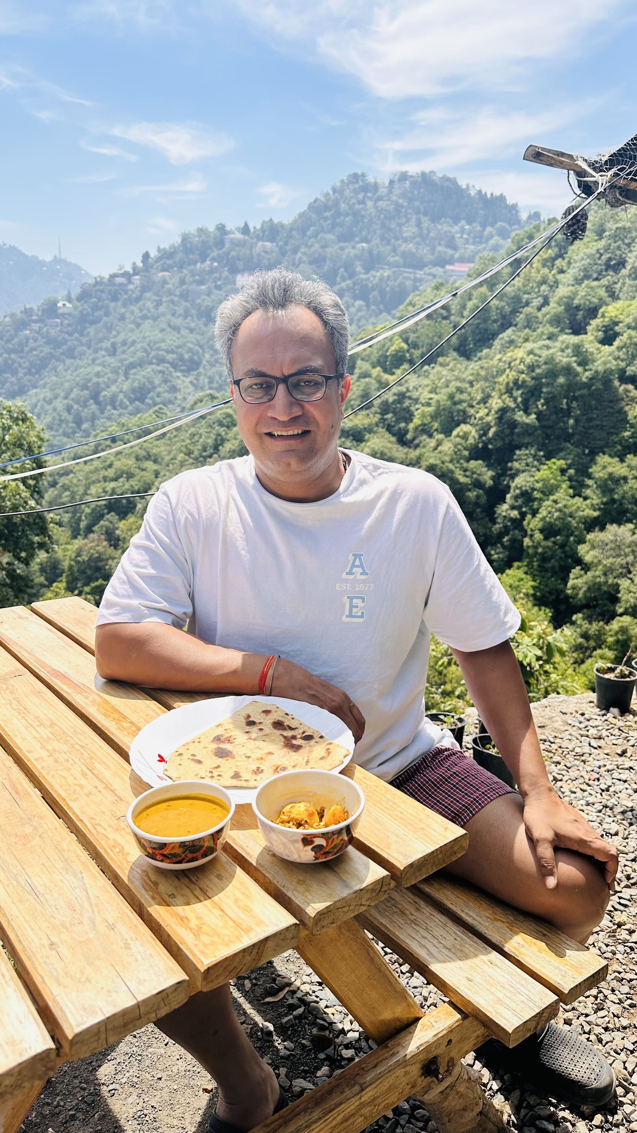 Una persona seduta a un tavolo da picnic in legno, con un piatto di cibo e due ciotole, sullo sfondo di un paesaggio montuoso.