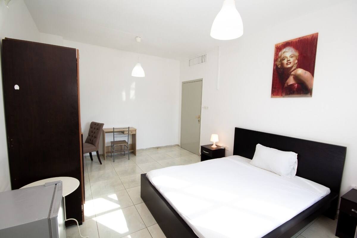 A bedroom with a bed, a desk, a chair, a nightstand, and a wall-mounted lamp.
