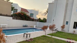 A swimming pool with a tiled deck, surrounded by a grassy lawn and small trees. - Villa 125  (Dubai)