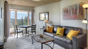 A living room with a leather sofa, a coffee table, and a dining area with chairs and a table. There is a painting on the wall and a view of trees through the window. - Pinnacle Place Ski-In/Out | Views, Fireplace, Covered Parking, Deck, Hot Tub (Durango)