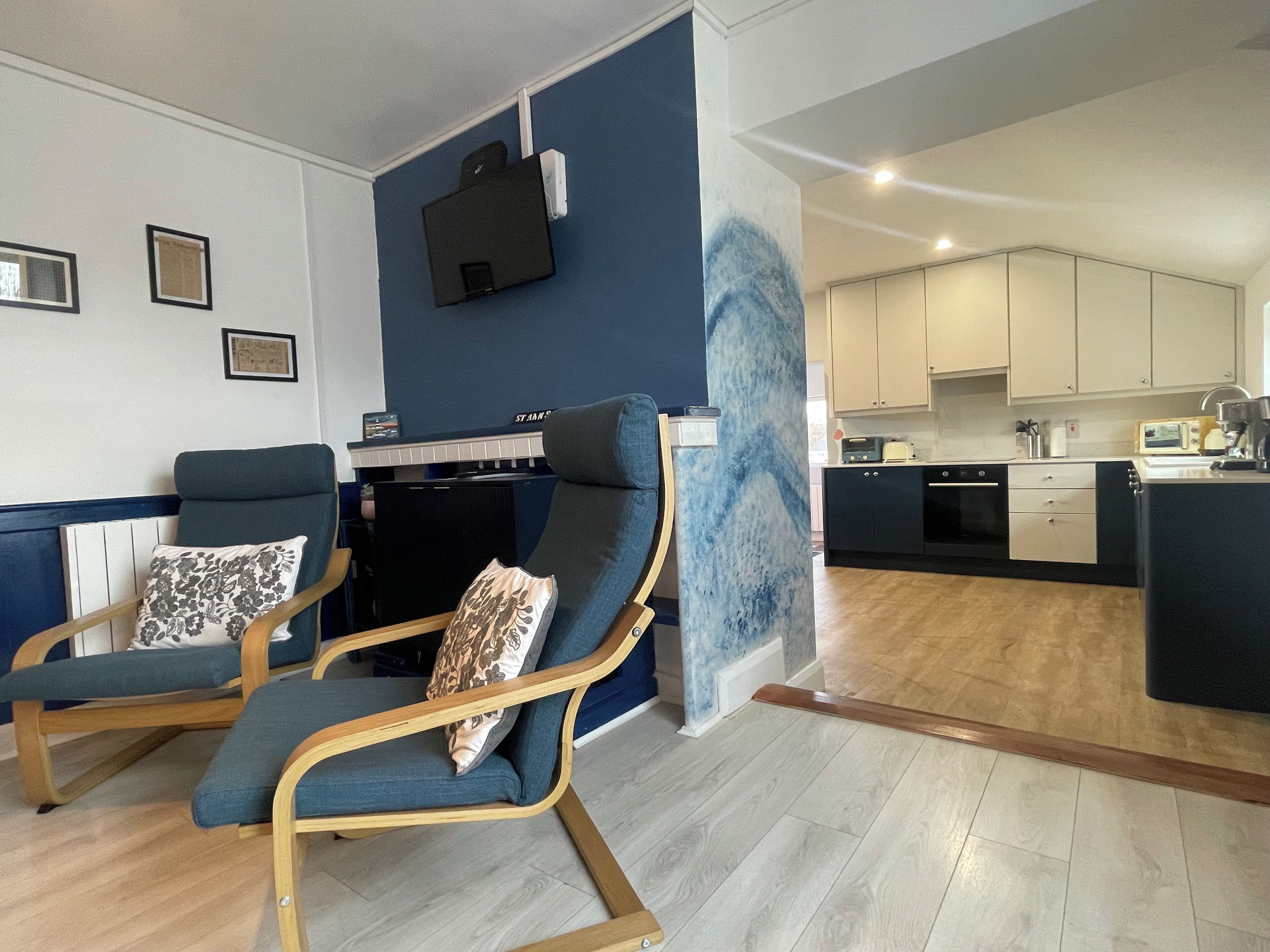 Una sala de estar moderna con dos sillones azules, una mesa de centro de madera y una pared azul de acento. Se ve una cocina al fondo, con armarios blancos y una estufa negra.