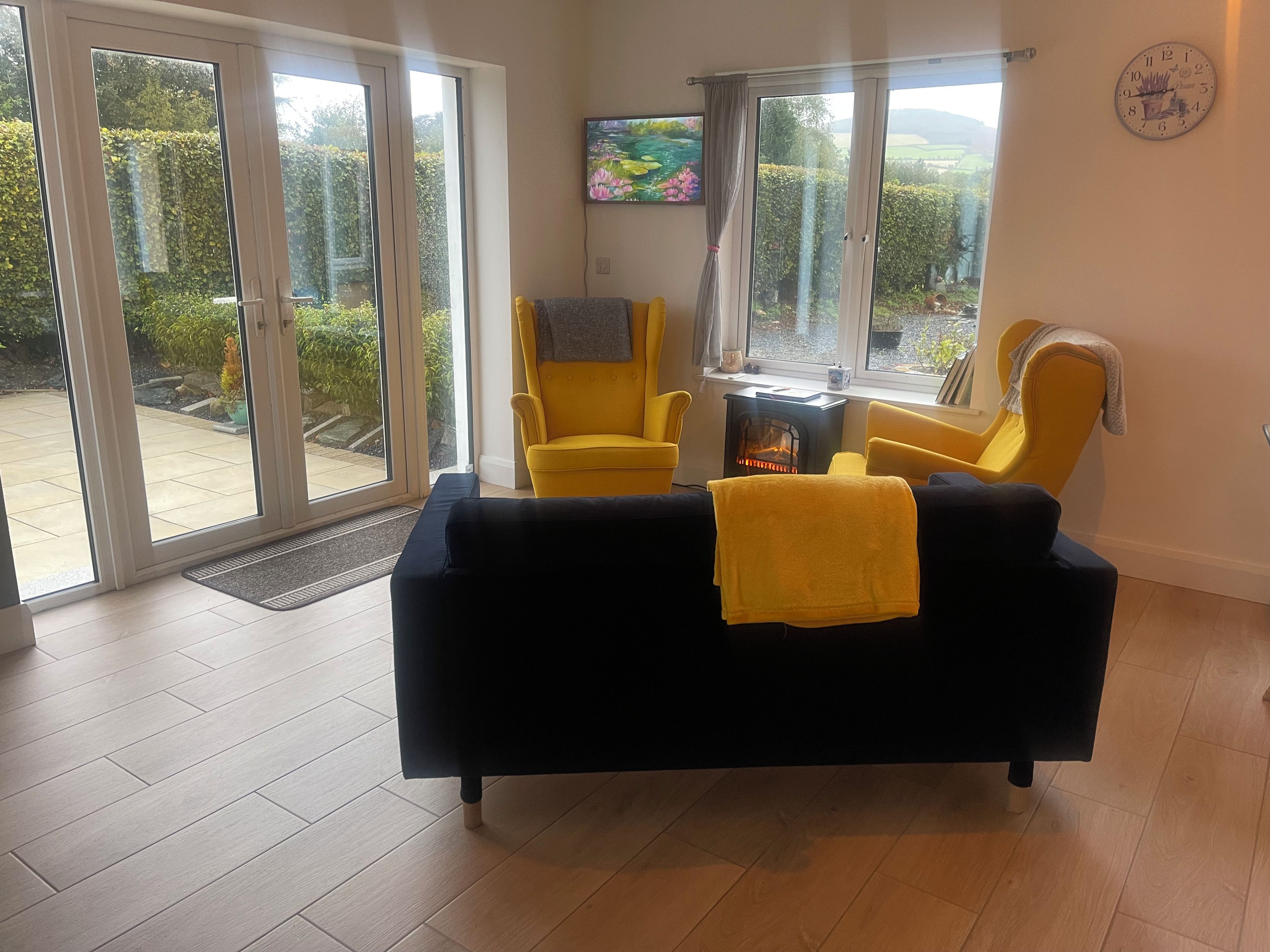 Una sala de estar con un sofá negro, sillones amarillos, una chimenea y puertas correderas de vidrio que dan a un jardín.
