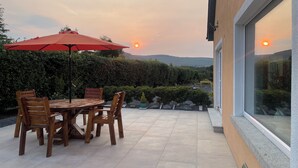 A patio with a round table and chairs, a red umbrella, and a view of the sunset over hills.