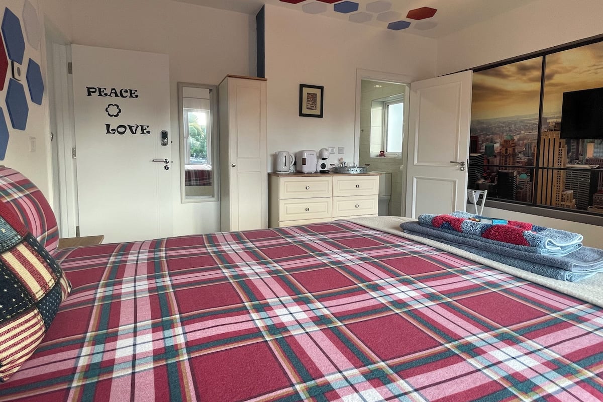 A bedroom with a plaid bedspread, a dresser with a mirror, a door with 'PEACE LOVE' decal, and a cityscape view through a window.