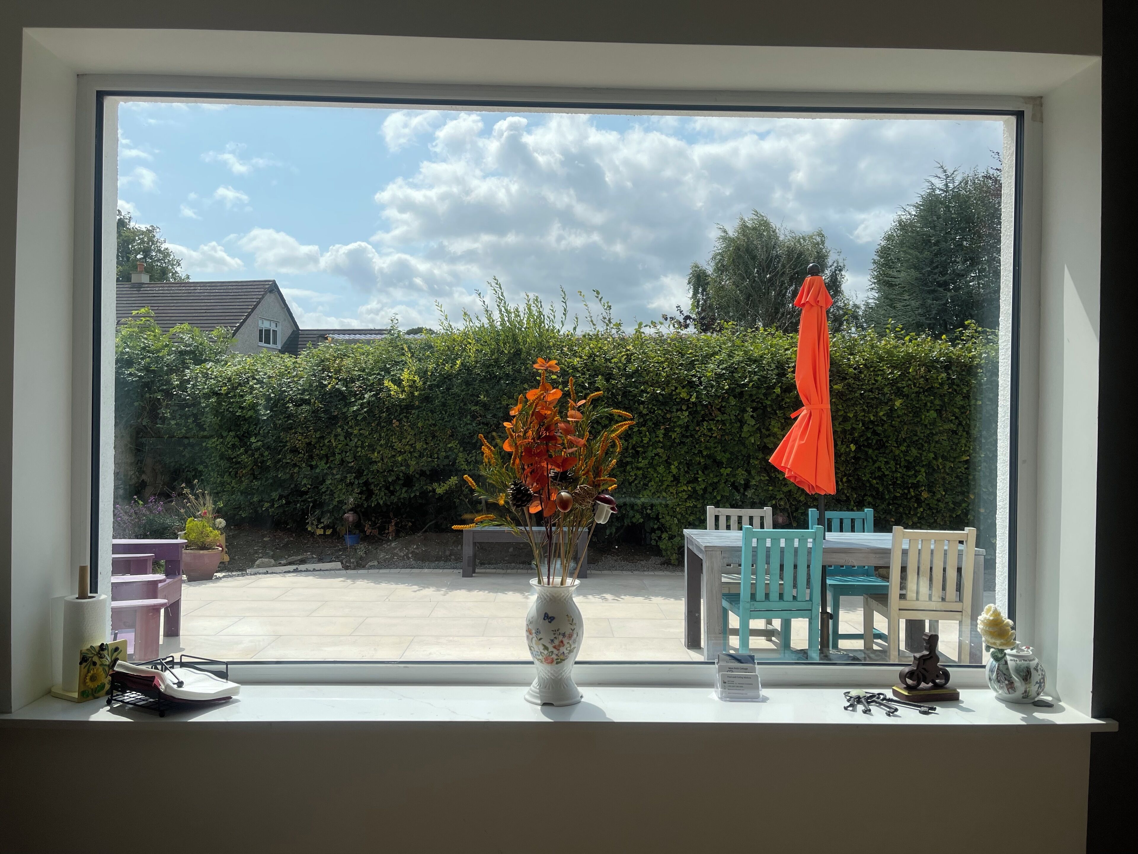 A window with a view of a patio area, including a table with chairs, a potted plant, and a red umbrella.