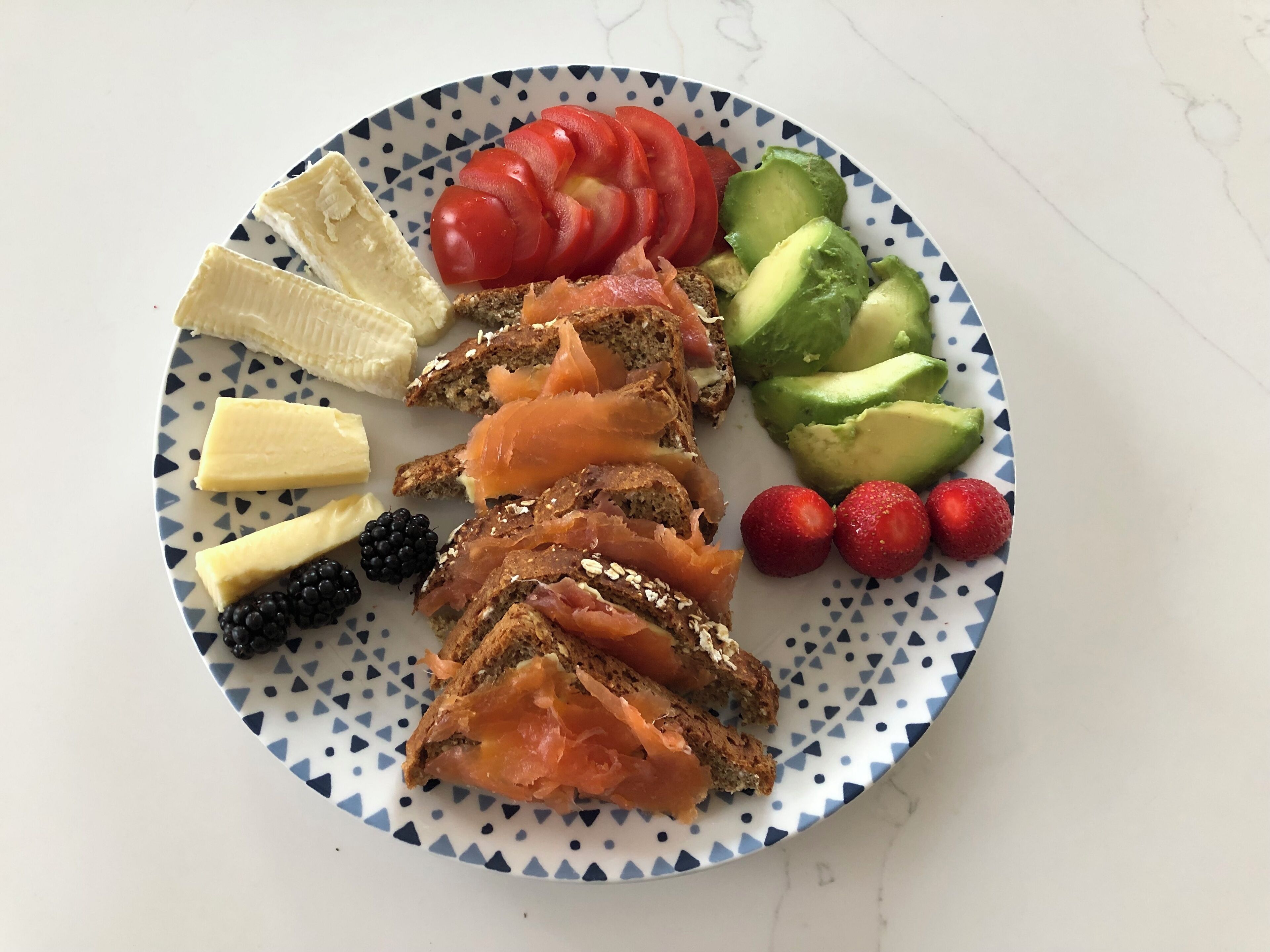 A plate with sliced salmon, avocado, strawberries, blackberries, cheese, and bread.