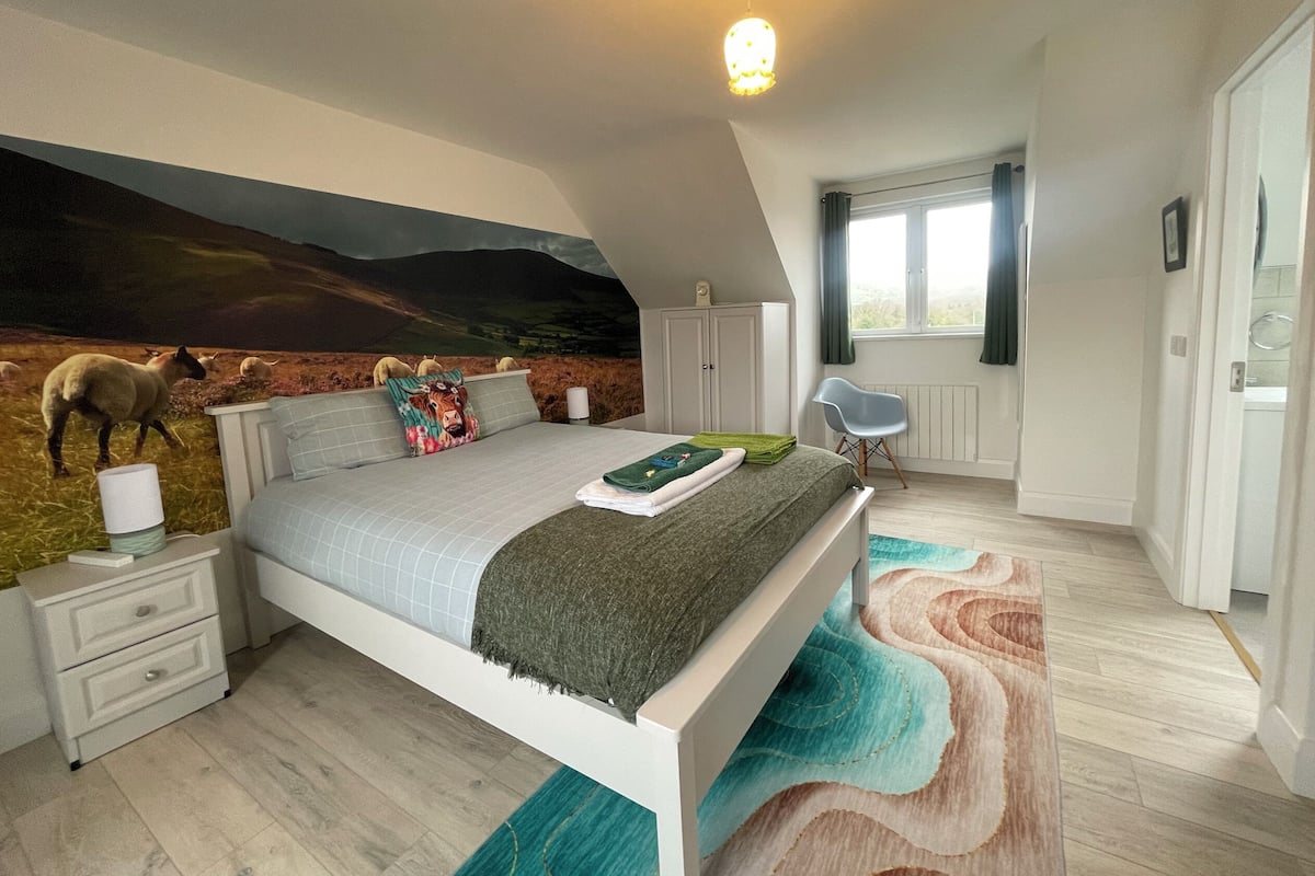 A bedroom with a mural of sheep, a bed with a green blanket, a white dresser, and a window with curtains.
