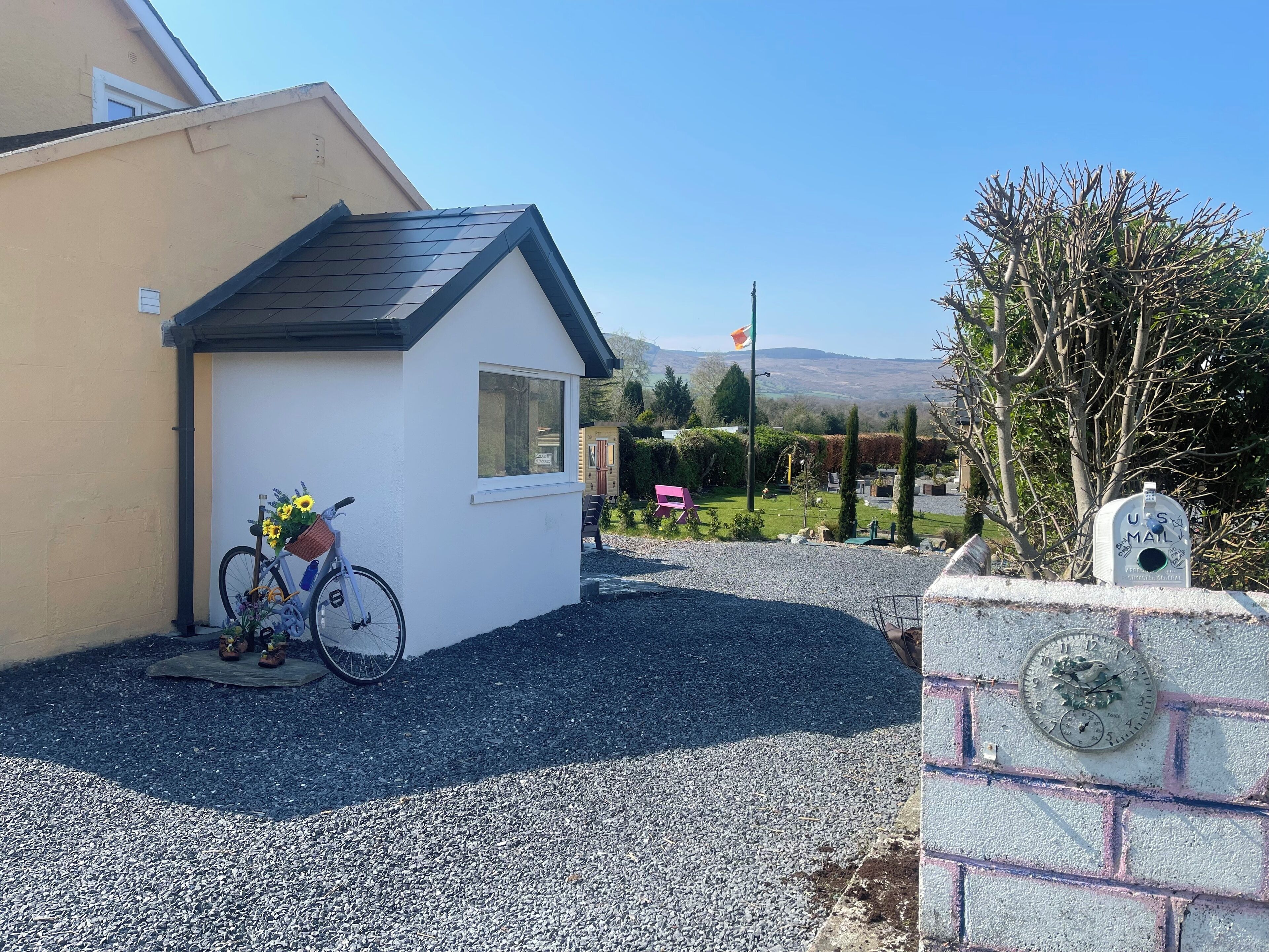 A small white building with a black roof, a bicycle with a basket of flowers, and a clock on a brick wall.
