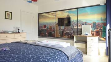 A hotel room with a large bed, a desk with a computer, a view of a cityscape, and a window with blinds.