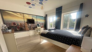 A bedroom with a cityscape view, a desk with a chair, a bed with a geometric-patterned bedspread, and a large window.