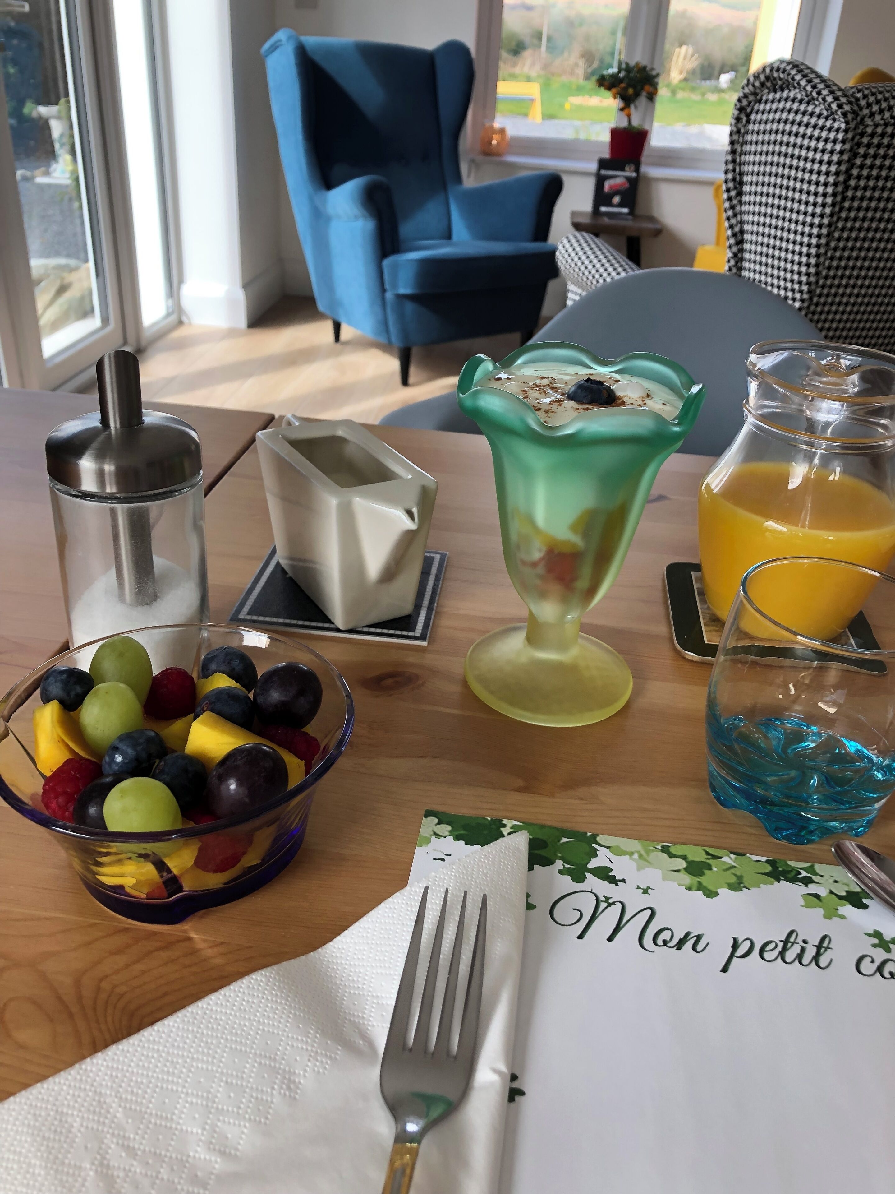 Una mesa de desayuno con un frutero, una jarra de vidrio verde, un vaso de jugo de naranja y una servilleta con un mensaje.