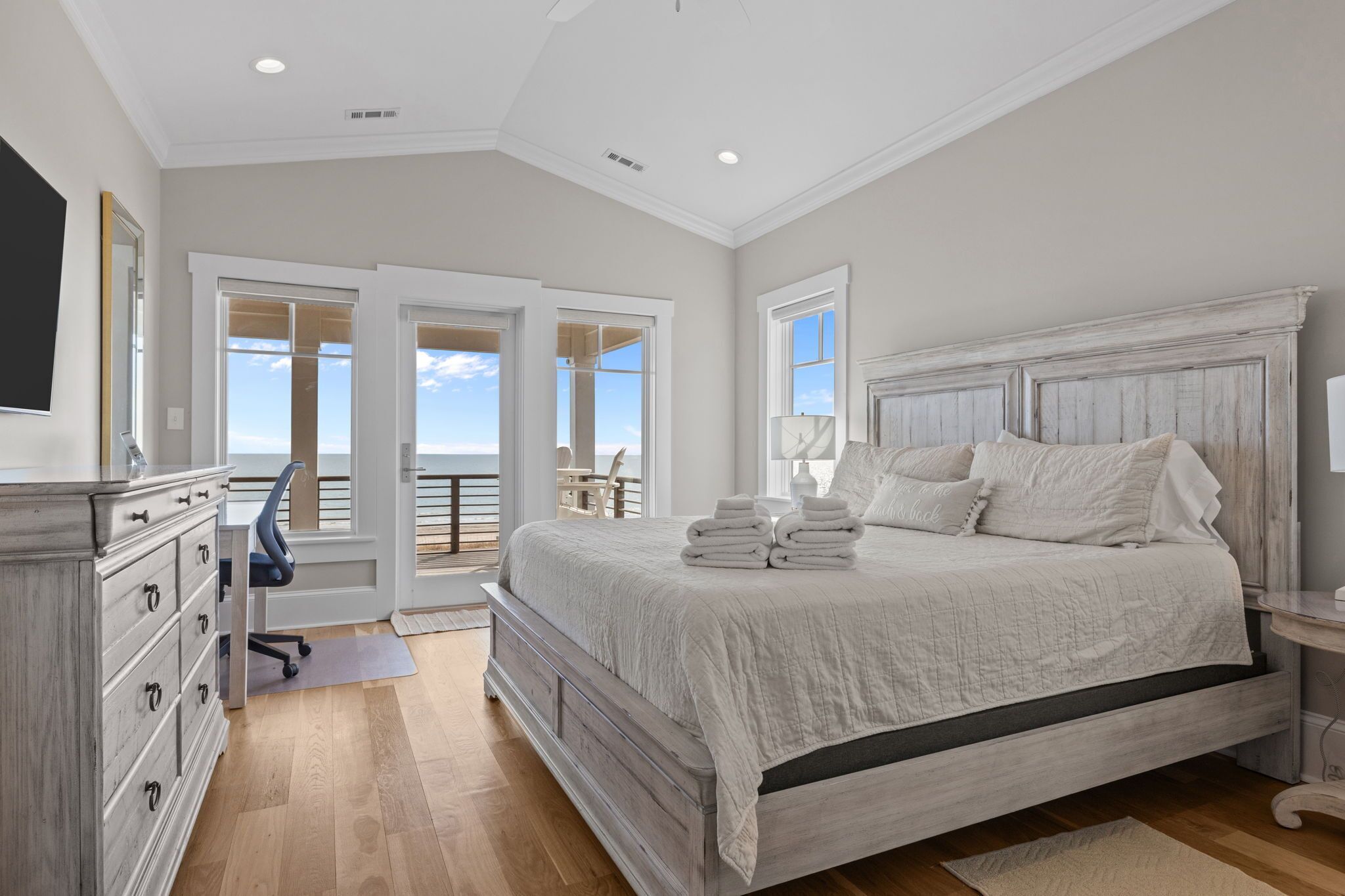 A bedroom with a large bed, a dresser, a desk, and a view of the ocean.