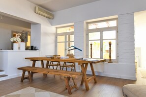 Un espace repas avec une table et des bancs en bois, des murs blancs et de grandes fenêtres.
