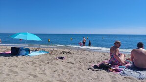 Une plage avec un parasol bleu, des gens dans l’eau et des vacanciers sur le sable.