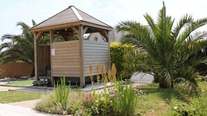 Un quiosco de madera con techo inclinado, rodeado de un jardín con diversas plantas y una palmera.
