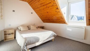 Un dormitorio con techo de madera inclinado, una cama con cubrecama gris, una mesita de noche, una pequeña estantería y una ventana con vistas al exterior.