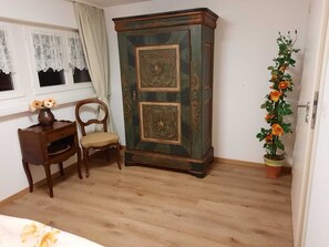 A room with a wooden floor, a green cabinet with ornate designs, a chair, a desk with a vase of flowers, and a potted plant with orange flowers. - Idyllic Farm 1 (Wildensbuch)