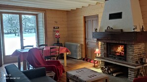 A cozy cabin interior with a stone fireplace, wooden walls, a dining area with a red tablecloth, and a view of a snowy landscape through large windows. - Authentic Jura farmhouse at the gateway to the Cascades du Hérisson (Saugeot)