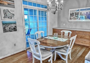 A dining area with a wooden table, white chairs, a chandelier, and a view through a glass door.