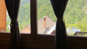 A window with curtains, revealing a mountainous landscape with snow-capped peaks and a green valley. - Chalet with nice view, garden and hot tub. Ideal for skiing and cycling! (Valloire)