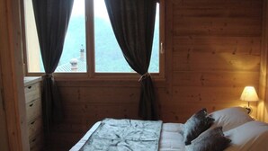 A wooden cabin bedroom with a bed, a nightstand with a lamp, and a window with curtains. - Chalet with nice view, garden and hot tub. Ideal for skiing and cycling! (Valloire)