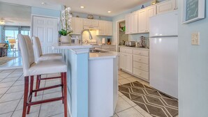 A modern kitchen with white cabinets, a central island with bar stools, a refrigerator, and a decorative wall sign. - Newly Remodeled Coastal Beachside Vacation Rental!! (Indian Rocks Beach)