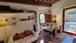 A cozy living room with a sofa, a dining table, and a TV. There are shelves with books and decorative items, and a window with a view of the outdoors.
