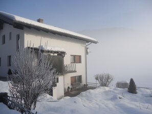 Une maison à deux étages avec un balcon, entourée d’un paysage enneigé et d’arbres.